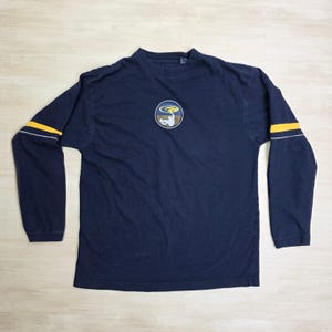 May include: Navy blue long-sleeved shirt with a crew neck. The shirt has a circular emblem on the chest and yellow and white stripes on the sleeves. The shirt is made of a soft, comfortable material.