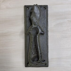 Peut inclure: Un heurtoir de porte en fonte de couleur gris foncé avec un design détaillé. Le heurtoir présente une forme de trou de serrure et est monté sur une plaque arrière rectangulaire avec deux trous. Conçu pour être monté sur une porte.
