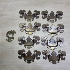 May include: Eight brass-toned metal drawer pulls with ornate, symmetrical backplates and swinging bail handles, accompanied by a small pile of matching metal mounting screws. The hardware features a weathered, aged finish with dark patina.