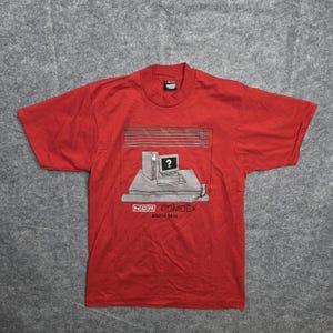 May include: Red t-shirt featuring a graphic of a computer with a question mark on the screen, set against a window with blinds. The text "NCR COMDEX BOOTH 1616" is printed below the graphic. The shirt has short sleeves and a crew neck.