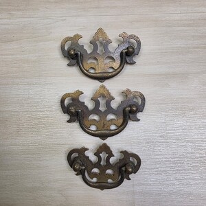 Puede incluir: Tres tiradores de cajón de latón de estilo antiguo con una pátina oscura y desgastada. Cada tirador presenta una placa trasera ornamentada y simétrica con detalles recortados intrincados y un asa curva móvil sujeta por dos perillas.