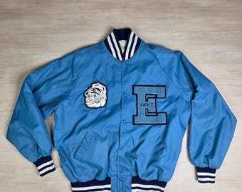 May include: Pale blue varsity jacket with navy and white striped detailing on the collar, cuffs, and hem. Includes a bulldog patch and a large letter "E" with "WAKE" stitched on the front. A classic athletic-style jacket.