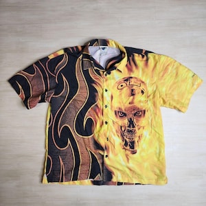 May include: A short-sleeved button-up shirt with a black and yellow print. The print features a skull with flames and a tribal design. The shirt has a button-up collar and short sleeves.