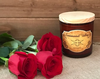 May include: A brown glass soy candle with a wooden lid. The candle is labeled "Sentimental Valley" and "Love Spell". Three red roses are in the foreground.