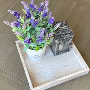 May include: A white wooden tray with a white ceramic pot with lavender flowers and the text "Merci Bouquet!" on the front. A black metal lantern with a glass window is on the tray.