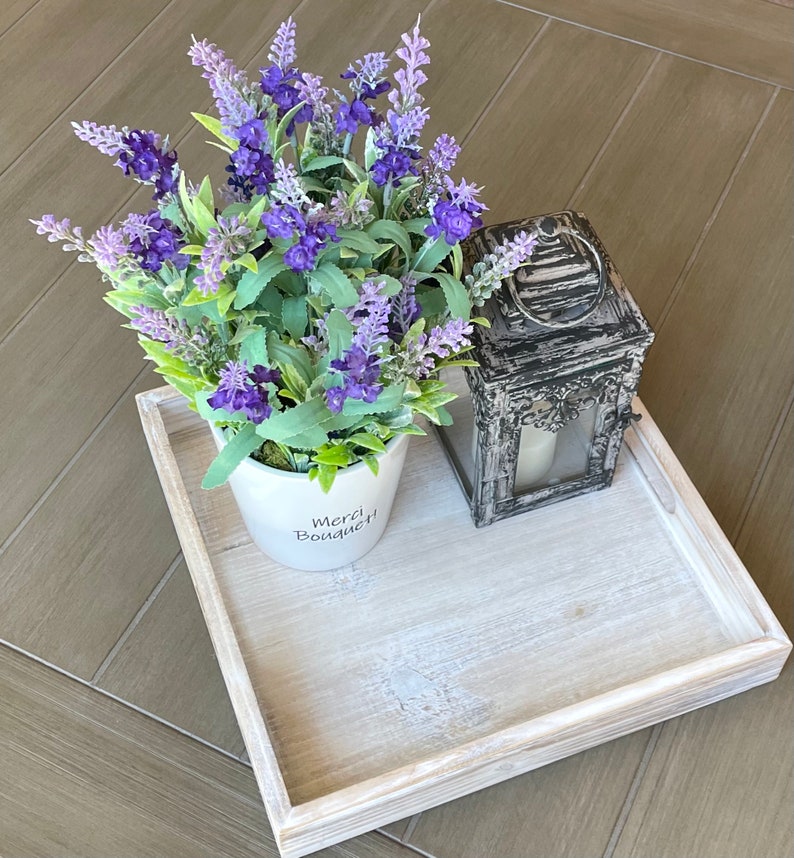May include: A white wooden tray with a white ceramic pot with lavender flowers and the text "Merci Bouquet!" on the front. A black metal lantern with a glass window is on the tray.