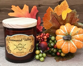 May include: A brown glass soy candle with a wooden lid. The candle has a label that says "Sentimental Valley Fall Festival Soy Candle". The candle is surrounded by fall-themed decorations, including leaves, berries, and a small pumpkin.