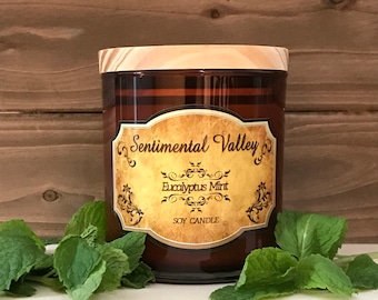 May include: A brown glass jar candle with a wooden lid. The label reads "Sentimental Valley" and "Eucalyptus Mint" with a decorative design. The candle is surrounded by green mint leaves.