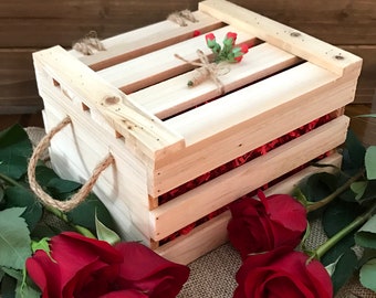 May include: A wooden crate with a natural finish and a jute rope handle. The crate is filled with red shredded paper and a small sprig of green leaves with red flowers. Three red roses are in the foreground on a burlap surface.