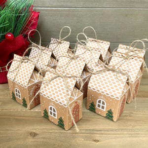May include: A collection of small, brown cardboard gift boxes designed to resemble gingerbread houses. Each box features a white roof, a red door, green trees, and a white window. The boxes are tied with twine and are arranged on a wooden surface.