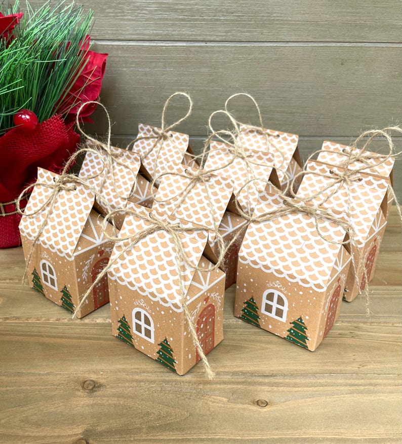 May include: A collection of small, brown cardboard gift boxes designed to resemble gingerbread houses. Each box features a white roof, a red door, green trees, and a white window. The boxes are tied with twine and are arranged on a wooden surface.