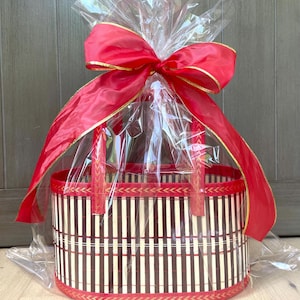 May include: A round bamboo gift basket with a red and gold ribbon bow. The basket has vertical brown and beige stripes, red trim, and handles. The basket is wrapped in clear cellophane.