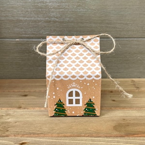 May include: A small, brown paper gift bag decorated with a gingerbread house design. The bag features a white scalloped roof, a white window, and two green Christmas trees. A twine bow secures the top.