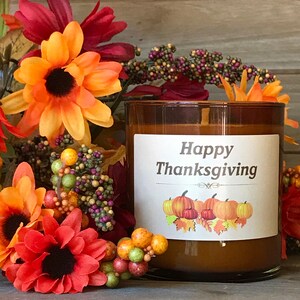 May include: A brown glass candle with a white label that reads "Happy Thanksgiving" with pumpkin illustrations. The candle is surrounded by orange, red, and yellow flowers and berries, creating a festive autumn theme.