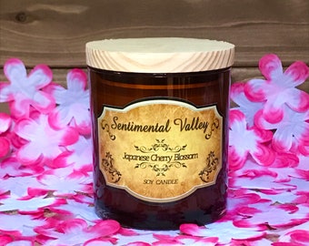 May include: A brown glass jar candle with a wooden lid. The candle is labeled "Sentimental Valley" and "Japanese Cherry Blossom" with a gold decorative border. The candle is surrounded by pink and white flower petals.