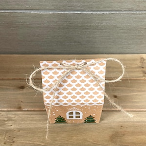 May include: A small, brown paper gift box with a white scalloped pattern and a twine bow. The box features a house design with a window and two green Christmas trees. The box is tied with natural twine.