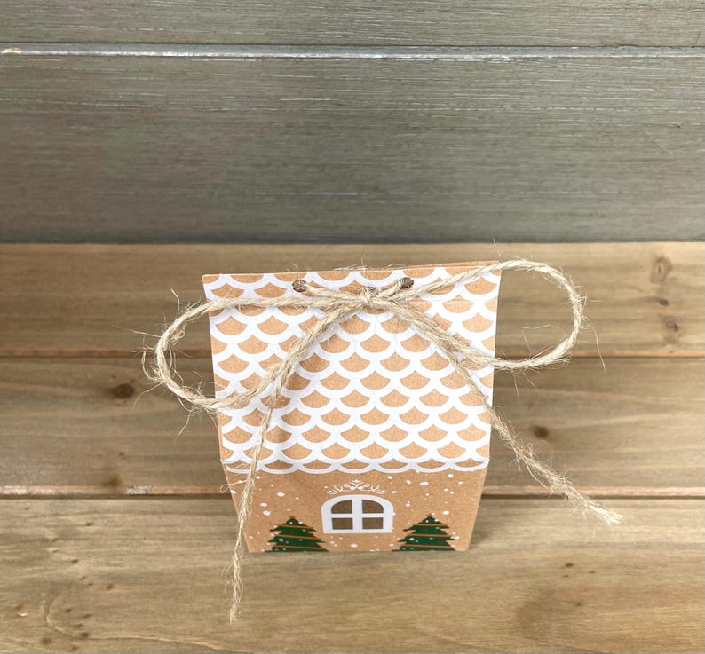May include: A small, brown paper gift box with a white scalloped pattern and a twine bow. The box features a house design with a window and two green Christmas trees. The box is tied with natural twine.
