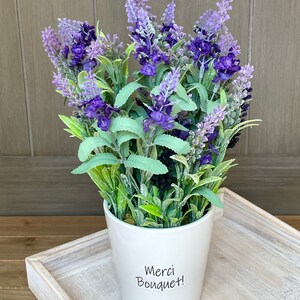May include: A white ceramic pot with a lavender bouquet. The pot has the text "Merci Bouquet!" written on it.