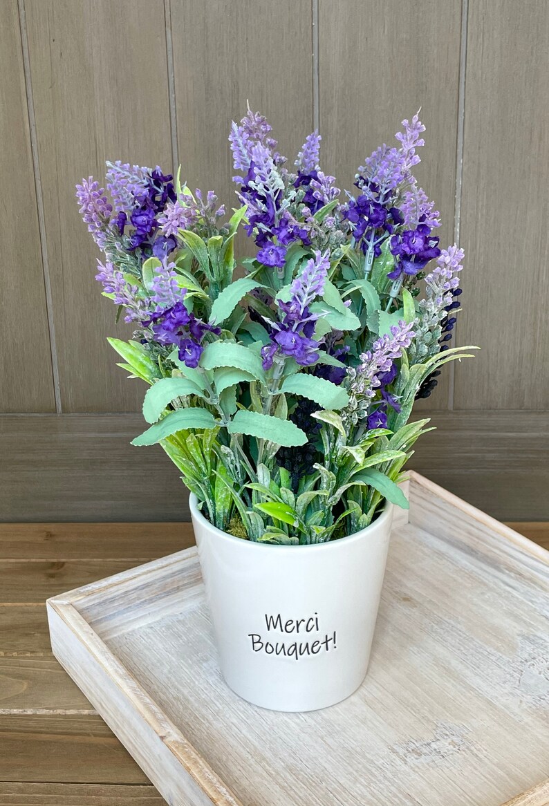 May include: A white ceramic pot with a lavender bouquet. The pot has the text "Merci Bouquet!" written on it.