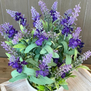May include: A white ceramic pot filled with a lush arrangement of purple lavender flowers. The flowers are in full bloom and have a delicate, fragrant scent.