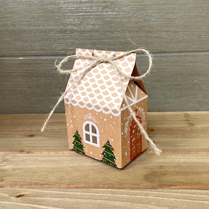 May include: A small, brown cardboard gift box shaped like a gingerbread house. The roof is white with a scalloped pattern, and the front features a white window, two green Christmas trees, and a red door. The box is tied with twine.