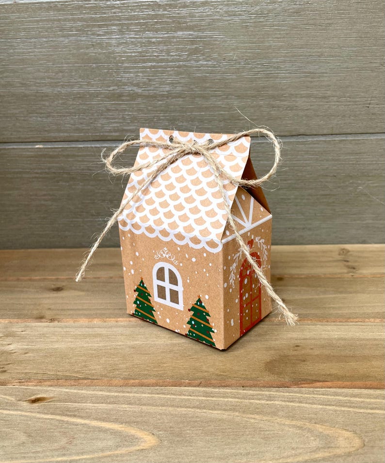 May include: A small, brown cardboard gift box shaped like a gingerbread house. The roof is white with a scalloped pattern, and the front features a white window, two green Christmas trees, and a red door. The box is tied with twine.