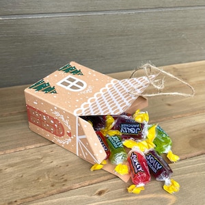 May include: A brown cardboard house-shaped box with a white window and green tree designs, spilling out colorful Jolly Rancher candies. The box is decorated with red dots and a white lace-like design. The candies are red, green, and purple.