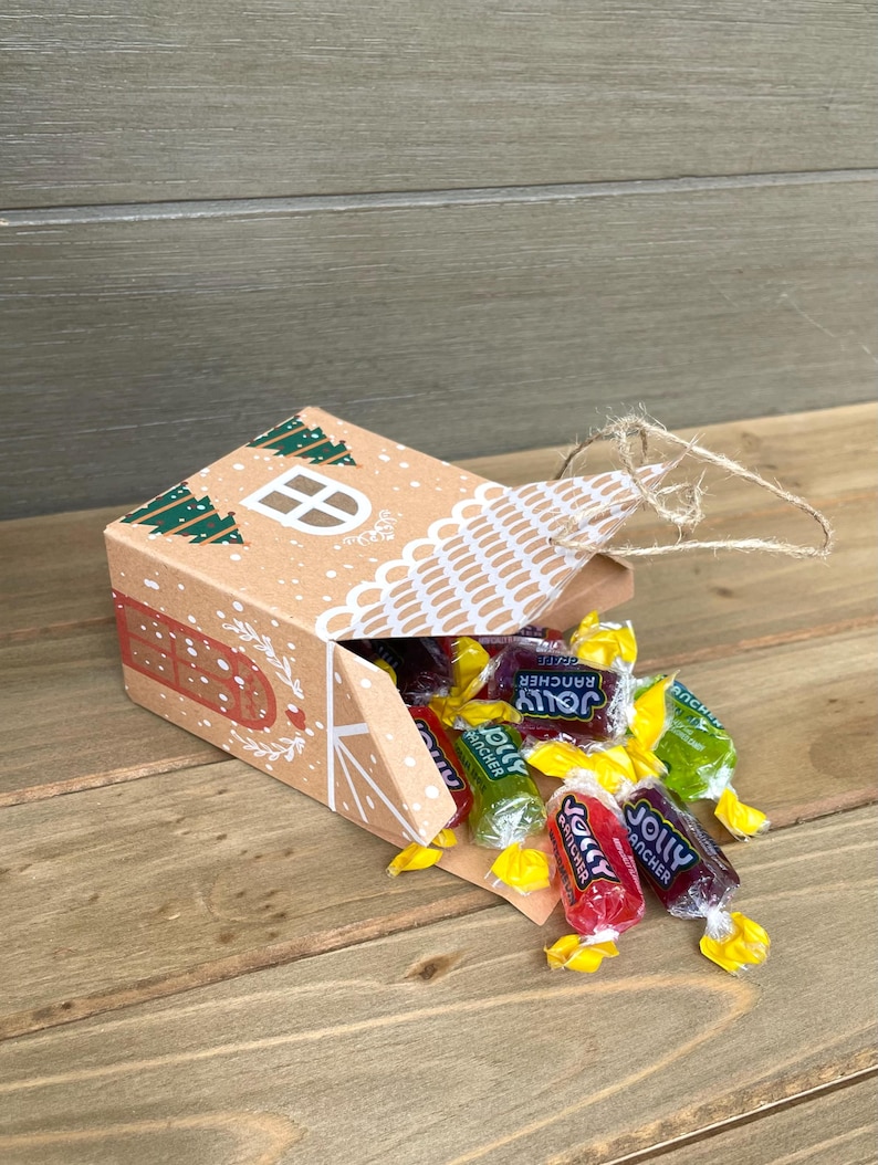 May include: A brown cardboard house-shaped box with a white window and green tree designs, spilling out colorful Jolly Rancher candies. The box is decorated with red dots and a white lace-like design. The candies are red, green, and purple.