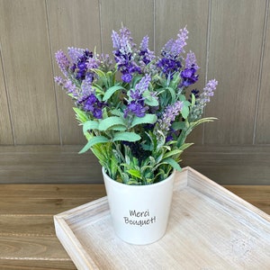 May include: A white ceramic pot with the text "Merci Bouquet!" on the front, filled with a bouquet of purple lavender flowers.