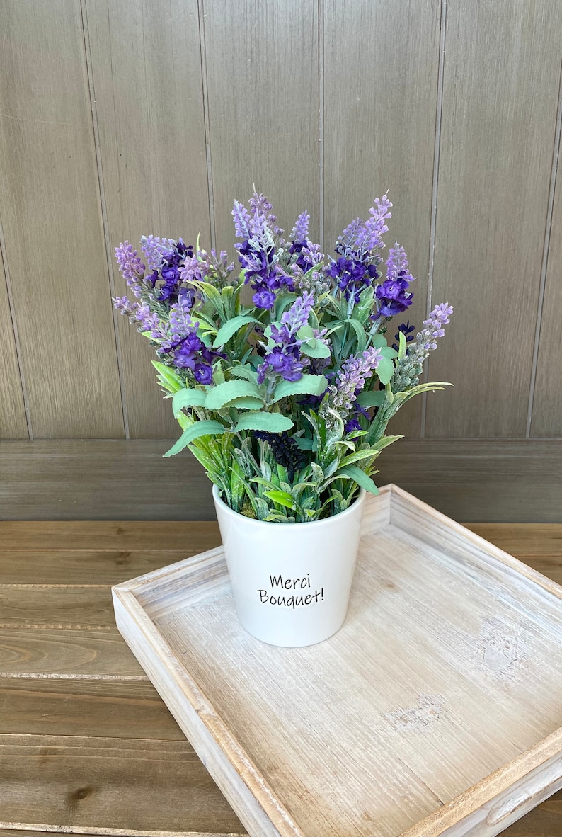 May include: A white ceramic pot with the text "Merci Bouquet!" on the front, filled with a bouquet of purple lavender flowers.