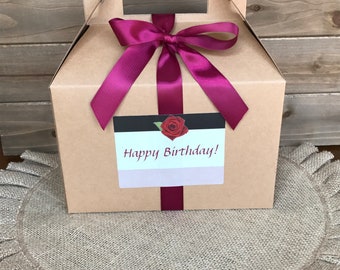 May include: A tan gift box with a handle, tied with a deep magenta satin ribbon and bow. A label with the words "Happy Birthday!" and a red rose is attached to the box. The box sits on a burlap placemat.