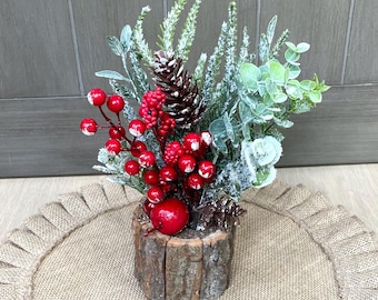 May include: A festive centerpiece featuring a wooden log container filled with faux greenery, red berries, and pinecones. The arrangement sits on a round burlap placemat, creating a rustic holiday display.