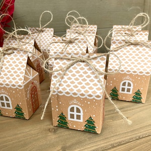 May include: Several small, brown paper gift boxes shaped like gingerbread houses, tied with twine. Each box features a white scalloped roof, a white window, and green Christmas trees. The boxes are arranged on a wooden surface.