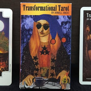 May include: A tarot card deck with three visible cards and a box. The box and cards feature illustrations of figures and the text "Transformational Tarot" and "Queen of Wands". The deck is on a dark surface.