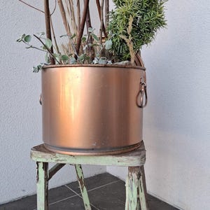 May include: A copper-colored metal planter with handles, filled with dried branches and greenery, sits on a distressed, light green wooden stool. The planter has a cylindrical shape and is placed against a white wall.