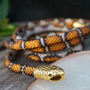 May include: A beaded snake necklace with a gold snake head charm. The necklace is made of black, white, and orange beads and is coiled on a wooden surface.