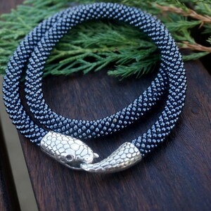 Pu&ograve; includere: Una collana di perline nere con una chiusura a serpente argentata. La collana &egrave; avvolta su una superficie di legno.