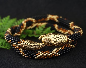Puede incluir: Un collar de serpiente de cuentas con un dije de cabeza de serpiente dorada. El collar es negro y dorado y está enrollado sobre una superficie negra.