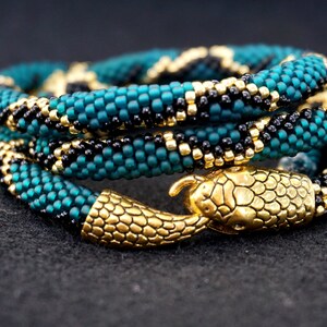 May include: A beaded bracelet with a snake design in teal and black colors. The bracelet has a gold snake head clasp.