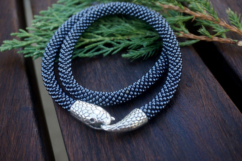 Pu&ograve; includere: Una collana di perline nere con una chiusura a serpente argentata. La collana &egrave; avvolta su una superficie di legno.