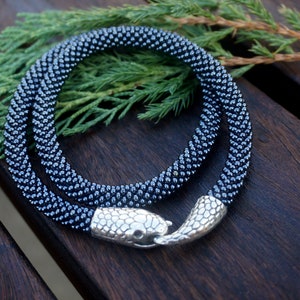 Pu&ograve; includere: Una collana di perline blu scuro con una chiusura a testa di serpente argentata. La collana &egrave; avvolta su una superficie di legno.