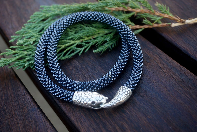 Pu&ograve; includere: Una collana di perline blu scuro con una chiusura a testa di serpente argentata. La collana &egrave; avvolta su una superficie di legno.