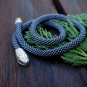 Pu&ograve; includere: Una collana di serpente in perle grigio scuro con un ciondolo a forma di testa di serpente argentato. La collana &egrave; avvolta su un ramo verde.