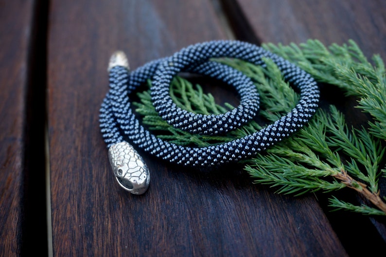 Pu&ograve; includere: Una collana di serpente in perle grigio scuro con un ciondolo a forma di testa di serpente argentato. La collana &egrave; avvolta su un ramo verde.