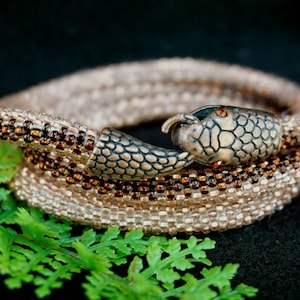 Peut inclure: Un bracelet en forme de serpent composé de perles, avec un fermoir en forme de tête de serpent en bronze. Le bracelet est fait de petites perles brun clair et brun foncé. Le fermoir en forme de tête de serpent est détaillé avec des écailles et a un œil rouge.