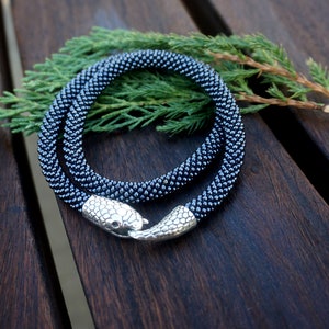 Pu&ograve; includere: Una collana di perline nere con una chiusura a forma di serpente. La collana &egrave; realizzata con piccole perline rotonde ed &egrave; avvolta su una superficie di legno.
