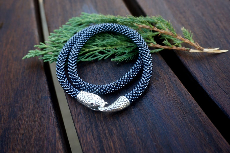 Pu&ograve; includere: Una collana di perline nere con una chiusura a forma di serpente. La collana &egrave; realizzata con piccole perline rotonde ed &egrave; avvolta su una superficie di legno.