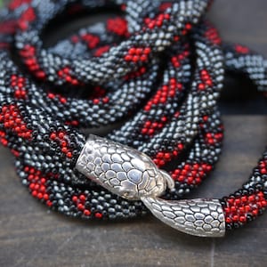 Puede incluir: Un collar de serpiente de cuentas con un cierre de cabeza de serpiente de plata. Las cuentas son negras, rojas y plateadas.