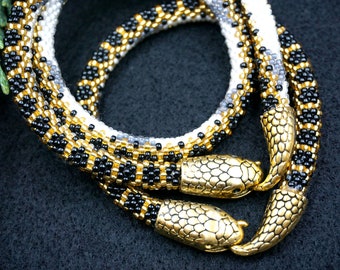 Puede incluir: Dos collares de serpiente de cuentas con cierres de cabeza de serpiente dorados. Los collares son negros, dorados y blancos.