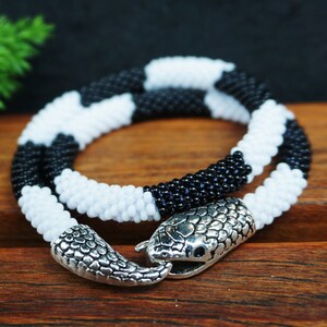 Puede incluir: Collar de cuentas negras y blancas con cierre de cabeza de serpiente plateado.